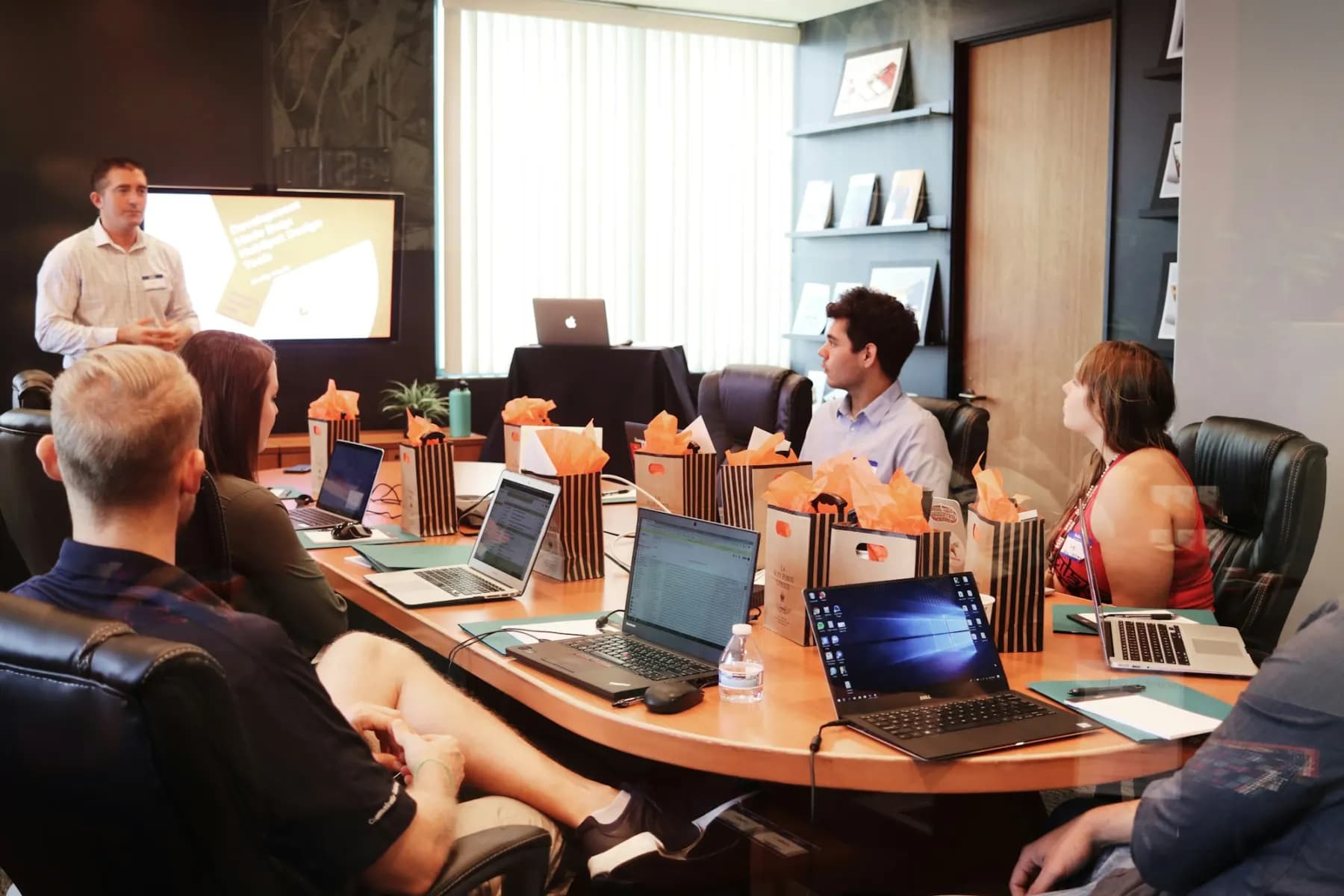 A team around a meeting table with laptops, one person standing at a screen to take them through a plan.