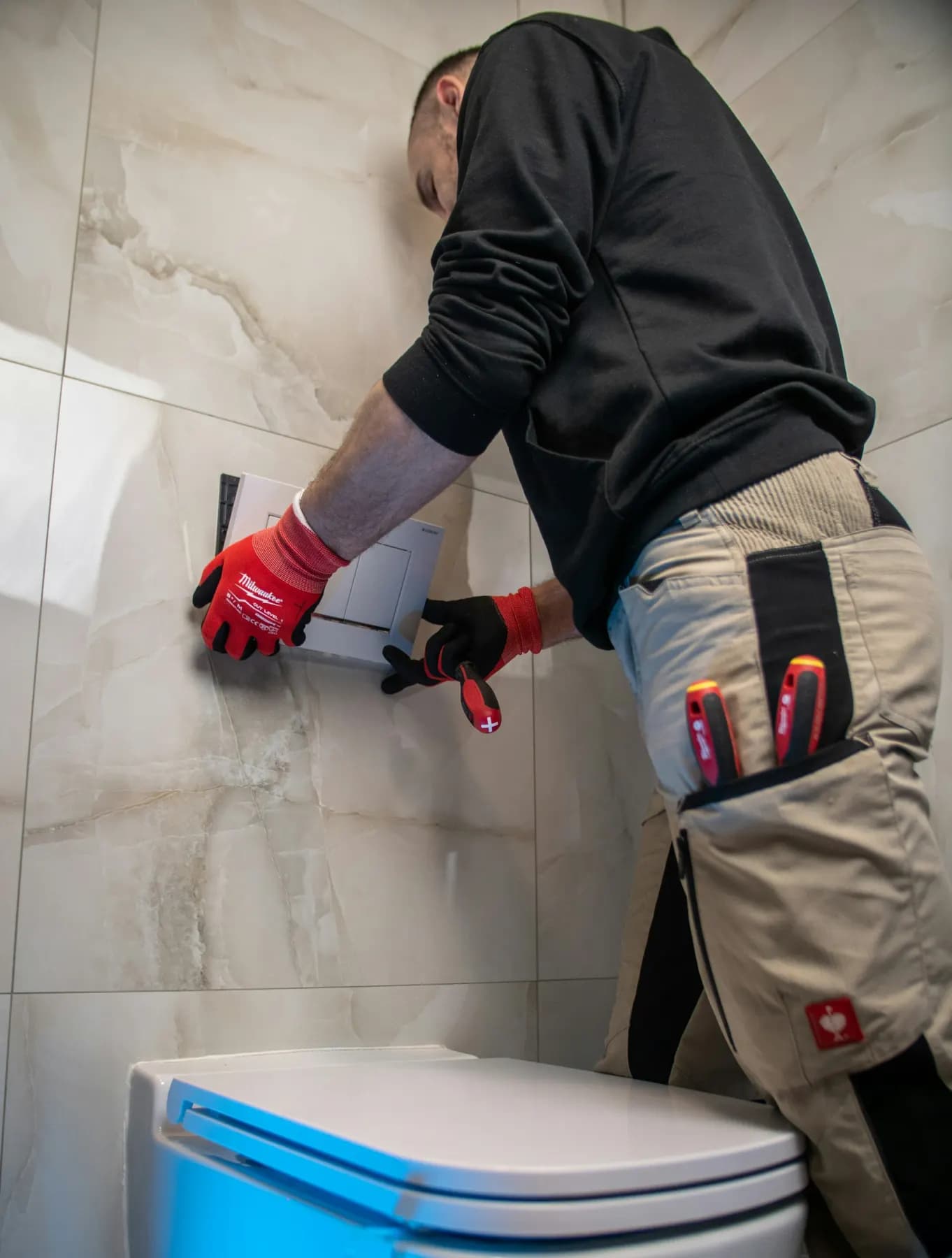A tradesman in work trousers and red gloves fitting a bathroom wall.